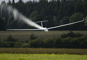 A modern glider crossing the finish line of a competition at high speed. It is jettisoning water that has been used as ballast.