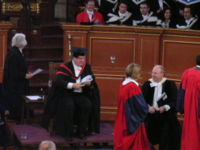 Degree ceremony at the University of Oxford. The Pro-Vice-Chancellor in MA gown and hood, Proctor in official dress and new Doctors of Philosophy in scarlet full dress. Behind them, a bedel, another Doctor and Bachelors of Arts and Medicine.